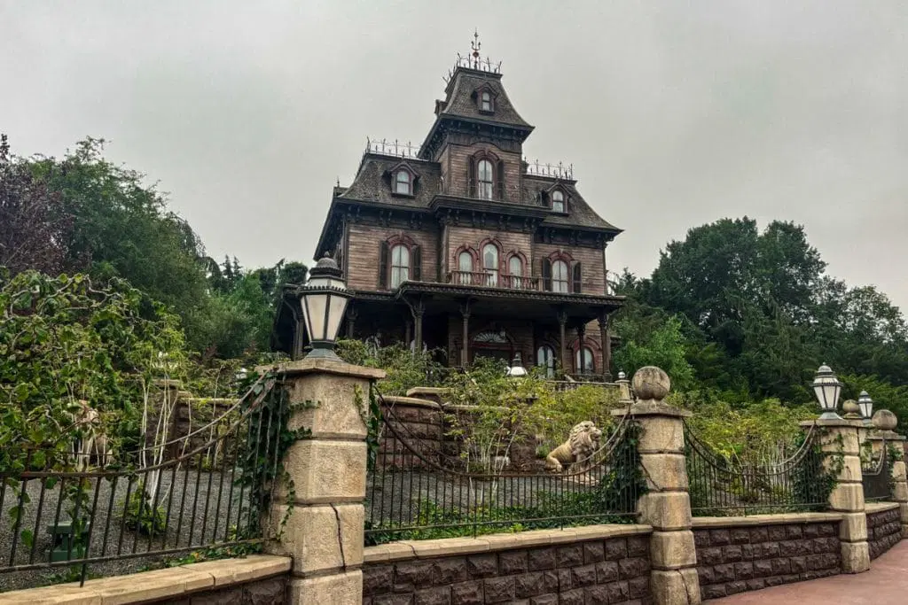 Phantom-Manor-1024x683.jpg A spooky, Victorian-style mansion surrounded by overgrown greenery, featuring intricate woodwork and multiple stories. The setting is eerie with an overcast sky, fitting the haunted theme of the Phantom Manor at Disneyland Paris.
