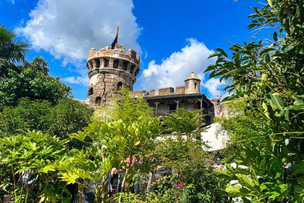 Pirates-of-the-Caribbean-1024x683.jpg A medieval-style stone tower partially hidden by lush green foliage and trees, set against a vibrant blue sky with white clouds. The structure gives a historic and adventurous vibe, likely part of a themed area in Disneyland Paris.