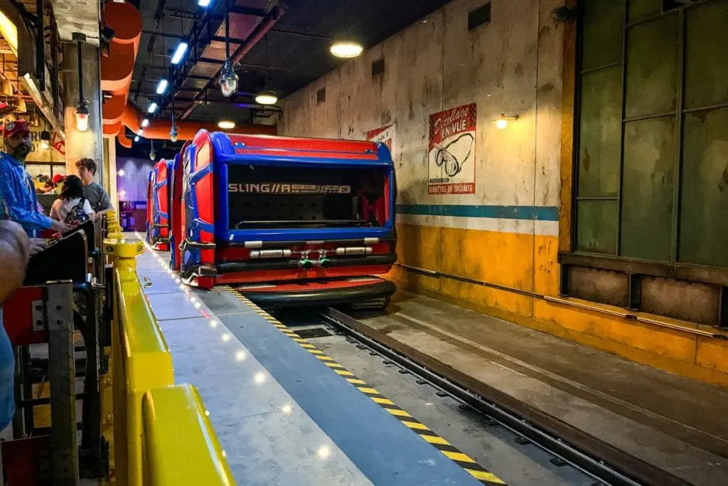 Spiderman-W.E.B-Adventure-1024x683.jpg A theme park ride vehicle designed to look like a futuristic, red and blue transport with the word 'SLING/RS' on the side. The ride is set indoors with a grungy, industrial theme. Guests are seen waiting in line, ready to board the ride.