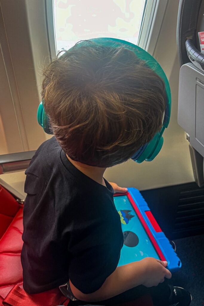 A young child wearing teal headphones watches a tablet screen while sitting in a red airplane seat next to a window, gazing outside mid-flight.