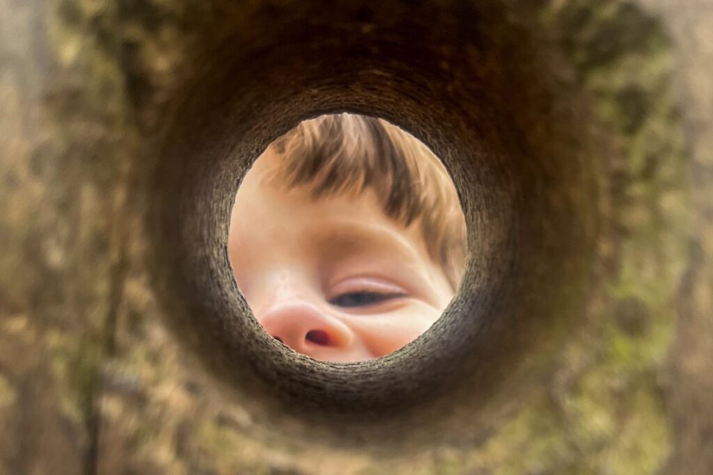 Mistakes Traveling with Kids A child’s face, partially visible and smiling, is seen through a round hole in a piece of wood, with soft lighting and a playful expression captured in the close-up view.