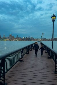 Two children walk hand in hand along a wooden pier at dusk, with street lamps glowing and a city skyline across the water in the background.