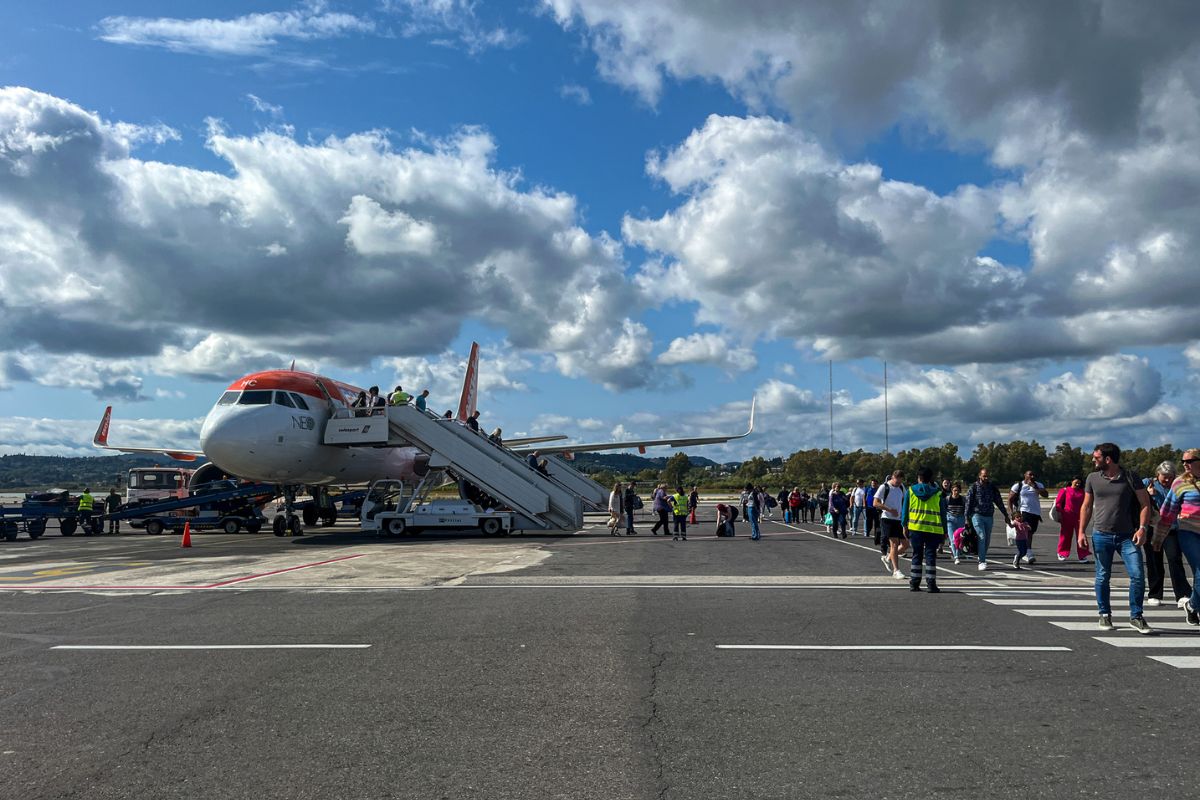 Passengers walk across the tarmac toward an EasyJet airplane, boarding via mobile stairs under a partly cloudy sky, with airport crew and luggage vehicles nearby.