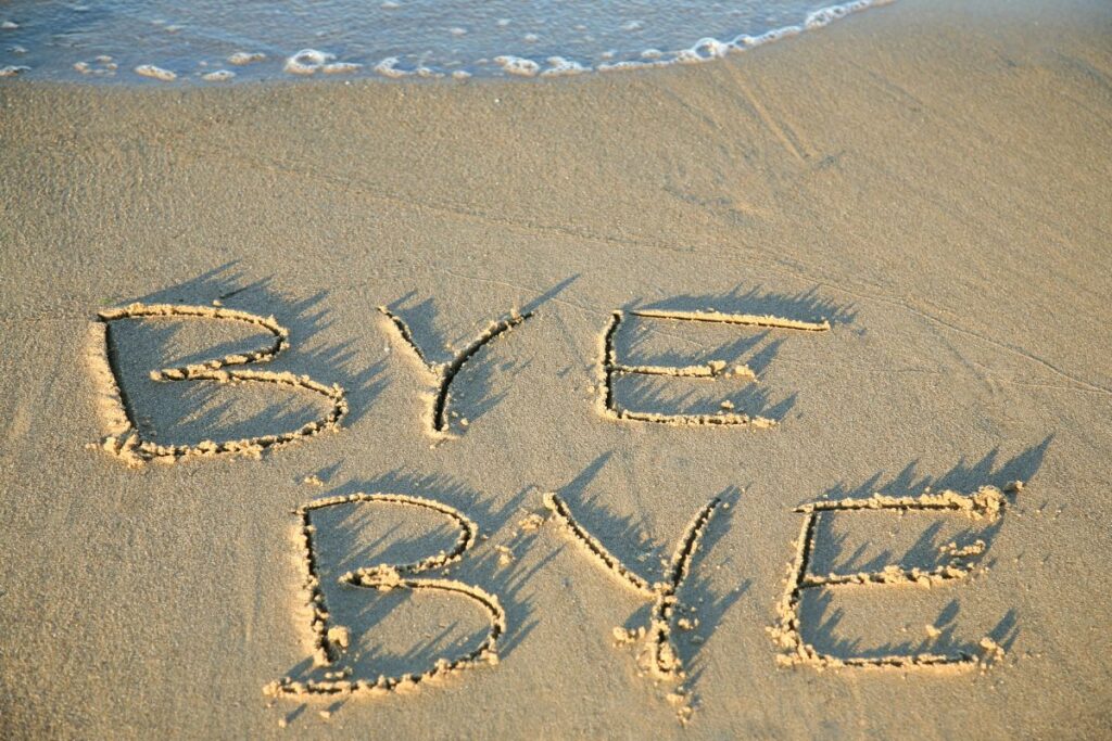 Bye Bye This image shows the words “BYE BYE” written by hand in smooth, damp sand at the edge of a beach. Each letter is carved deeply enough to create small ridges of sand along the edges, casting soft shadows in the low, warm light. Gentle waves approach from the top of the image, with a thin line of white foam creeping closer to the writing, suggesting the message may soon be washed away. The sand appears golden and evenly textured, with no footprints nearby, creating a calm and uncluttered scene. The positioning of the text near the waterline emphasizes the temporary nature of the message. The image conveys a sense of closure, transition, or farewell, paired with the peaceful rhythm of the sea and the passage of time.