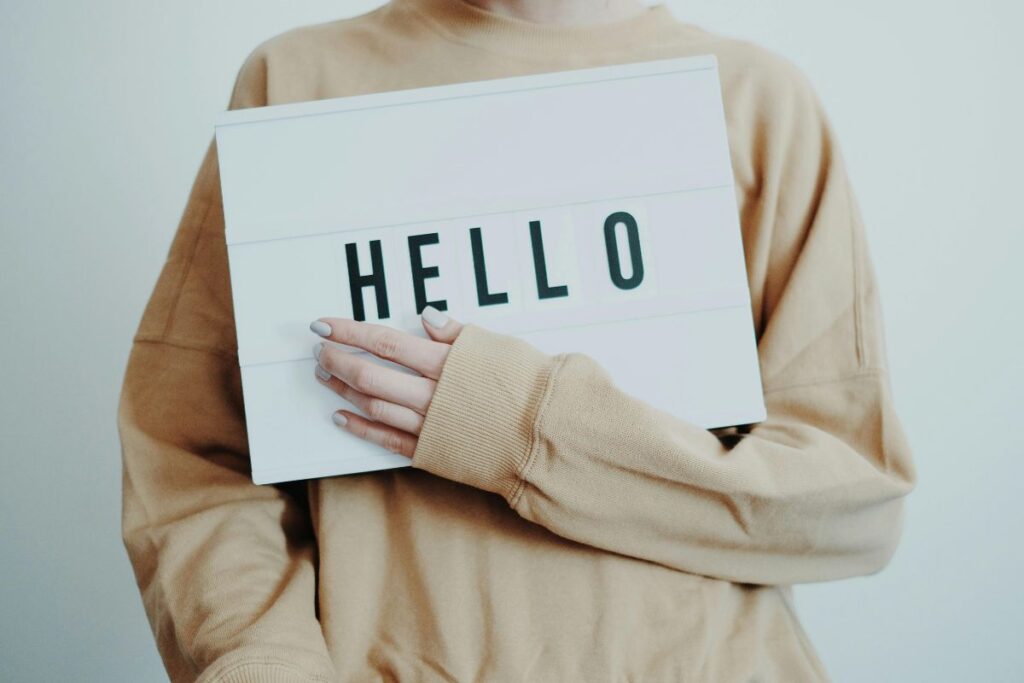 The image shows the upper body of a person standing against a plain, light-colored background while holding a rectangular white lightbox sign with the word “HELLO” displayed in bold, black capital letters. The person is wearing a loose, beige or tan sweatshirt with long sleeves that partially cover their hands, creating a relaxed and casual appearance. One hand rests gently across the front of the sign, with neatly groomed nails visible. The person’s face is cropped out of the frame, keeping the focus on the message rather than the individual. The clean background and neutral clothing tones draw attention to the word “HELLO,” making it the clear focal point of the image. The overall composition feels minimal, welcoming, and modern, suggesting an introduction, greeting, or starting point for content.