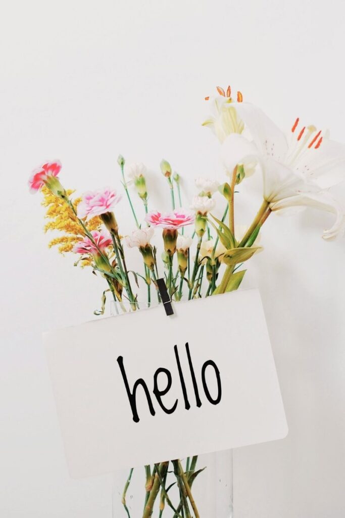 This image features a simple glass vase filled with a small bouquet of flowers, placed against a bright white background. The arrangement includes delicate pink blossoms, white lilies, and hints of yellow foliage, creating a soft and cheerful color palette. Attached to the stems near the front is a small white card clipped in place, displaying the handwritten word “hello” in lowercase black letters. The flowers rise upward, some leaning slightly, giving the arrangement a natural, unstructured look. The bright lighting and uncluttered background create an airy, fresh atmosphere. The combination of flowers and the greeting card conveys warmth, friendliness, and a gentle sense of welcome. The image feels calm and inviting, often associated with introductions, new beginnings, or thoughtful gestures.