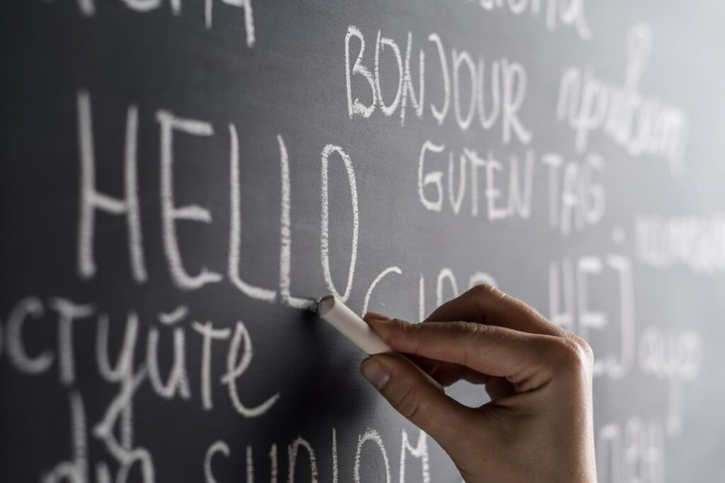 The image shows a close-up of a black chalkboard covered with handwritten greetings in different languages. A hand holding a piece of white chalk is captured mid-motion while writing the word “HELLO” in large, slightly textured letters. Surrounding the central word are other greetings written in chalk, including variations such as “bonjour” and “guten tag,” arranged in different sizes and orientations across the board. The chalk markings appear slightly smudged in places, emphasizing the tactile, handwritten nature of the scene. The shallow depth of field keeps the writing hand and the word “HELLO” in sharp focus while the surrounding text fades softly into the background. The image conveys communication, language learning, and inclusivity, highlighting how a simple greeting can connect people across cultures.