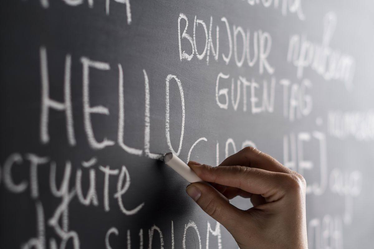 The image shows a close-up of a black chalkboard covered with handwritten greetings in different languages. A hand holding a piece of white chalk is captured mid-motion while writing the word “HELLO” in large, slightly textured letters. Surrounding the central word are other greetings written in chalk, including variations such as “bonjour” and “guten tag,” arranged in different sizes and orientations across the board. The chalk markings appear slightly smudged in places, emphasizing the tactile, handwritten nature of the scene. The shallow depth of field keeps the writing hand and the word “HELLO” in sharp focus while the surrounding text fades softly into the background. The image conveys communication, language learning, and inclusivity, highlighting how a simple greeting can connect people across cultures.