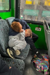 Two children sleep curled together on a bus seat, leaning against each other while traveling on a rainy day.