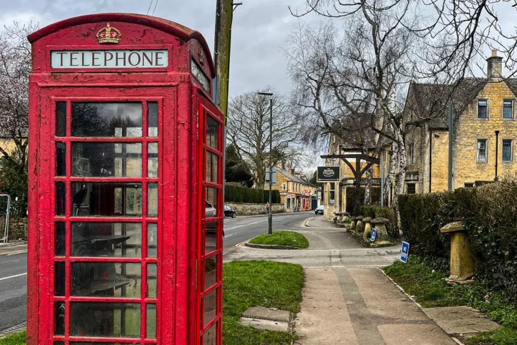 The image shows a classic red British telephone box standing on a village sidewalk beside a quiet road. The phone box is weathered, with visible chips and texture in the red paint, and the word “TELEPHONE” displayed at the top beneath a small crown emblem. It sits near a patch of grass and a paved footpath that curves gently into the distance. In the background, stone buildings with pitched roofs line the street, suggesting a traditional English village setting. Bare trees frame the scene, indicating late autumn or early spring. The road is lightly trafficked, with no visible crowds, giving the image a calm, timeless feel. The bold red of the phone box contrasts strongly with the muted stone, grey sky, and earthy tones of the surroundings, making it the clear focal point.