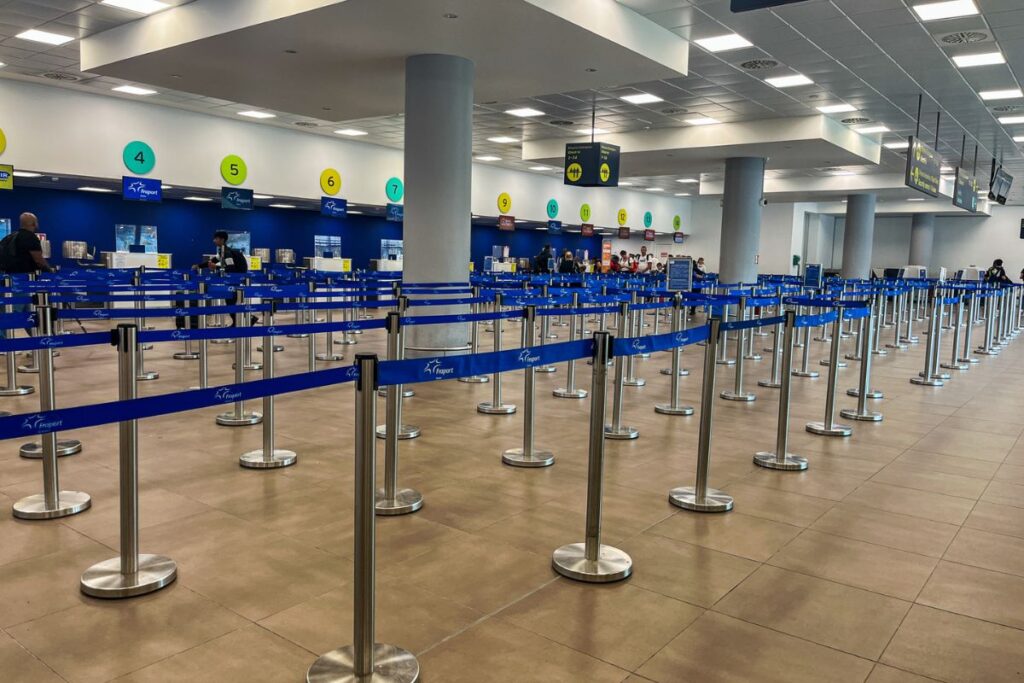 This image captures a wide view of an airport check-in hall that appears calm and largely empty. The foreground is filled with long rows of metal queue barriers connected by blue fabric straps, creating a zigzag path for passengers waiting to check in. The barriers stretch across the polished floor, emphasizing the scale and organization of the space. In the background, check-in counters line the wall, each marked with large, circular number signs in bright colors such as green, yellow, and blue. A few airport staff and travelers are scattered near the counters, but there are no visible crowds. Tall gray columns support the ceiling, which is fitted with evenly spaced lights that illuminate the hall with a neutral, even glow. Overhead signs hang from the ceiling, directing passengers to different services and areas. The overall scene feels orderly, quiet, and spacious, suggesting an early hour or a less busy travel period, and highlighting the structured process travelers follow before passing through security.