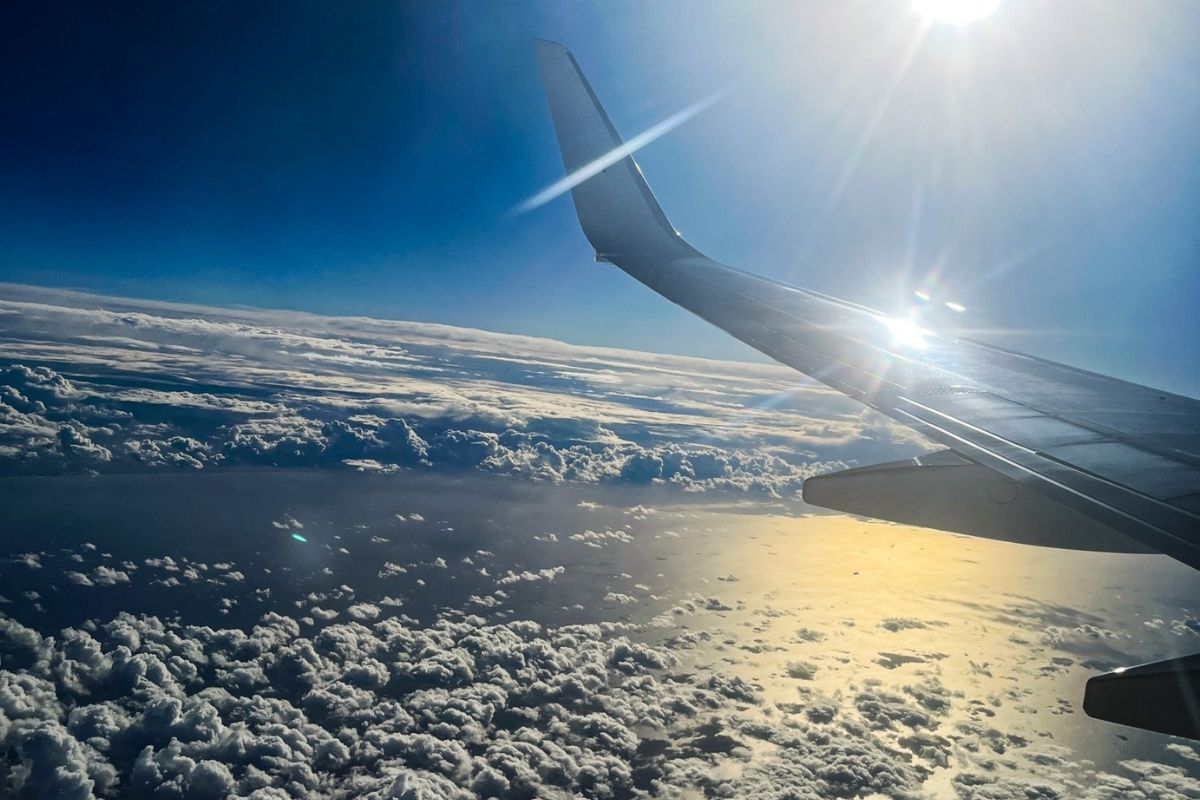 Sunlight reflects off an airplane wing above a layer of clouds, with blue sky and ocean visible far below during a daytime flight.