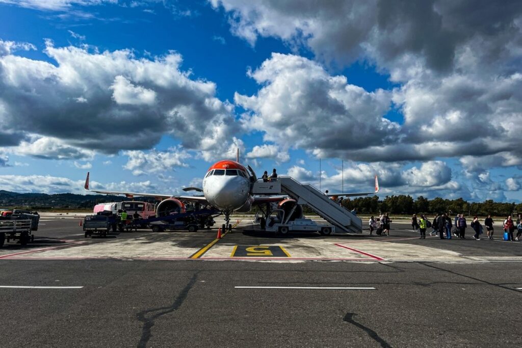 The image shows an airplane parked on an airport runway under a dramatic sky filled with large, billowing clouds. Passengers are boarding the aircraft using a set of portable stairs positioned at the front door. People stand in a loose line on the tarmac, some carrying small bags or backpacks. Ground crew vehicles and luggage carts are visible to the left of the plane, indicating active airport operations. The airplane’s nose faces the camera, with its engines and wings extending outward on either side. The contrast between the dark runway, bright white aircraft, and vivid blue sky creates a striking scene. The image captures the transition moment of travel, evoking anticipation, movement, and the practical realities of flying, particularly in smaller or regional airports.