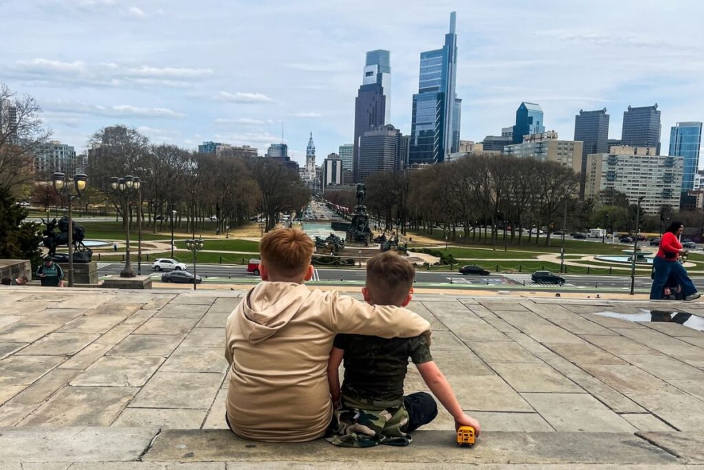 Kids Philadelphia The image shows two children sitting side by side on wide stone steps, viewed from behind. One child has an arm wrapped around the other’s shoulders in a gentle, affectionate gesture. In front of them stretches a large urban park with pathways, trees, and a central monument. Beyond the park, a city skyline rises, featuring tall modern buildings and historic architecture in the distance. Cars move along the road below, and pedestrians are scattered throughout the scene. One child holds a small toy vehicle near their side, adding a quiet, playful detail. The image conveys a reflective pause during sightseeing, emphasizing companionship, scale, and the contrast between childhood closeness and the vast cityscape ahead.