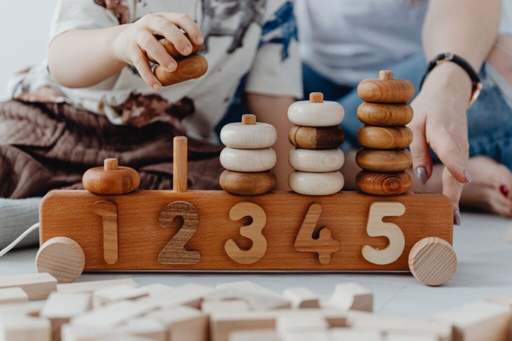 The image shows a child’s hands interacting with a wooden counting toy placed on the floor. The toy features carved numbers one through five along its base, with stacked wooden rings arranged vertically above each number. The rings vary in color from light natural wood to darker brown, highlighting contrast and progression. A child lifts one ring while an adult’s hand rests nearby, suggesting guided play or shared learning. Additional wooden blocks are scattered on the floor in the foreground, reinforcing the play-based setting. The materials appear smooth and natural, emphasizing tactile exploration. The scene conveys early numeracy learning through hands-on activity, focusing on counting, sorting, and fine motor skills. The environment feels calm, supportive, and educational, illustrating how numbers can be introduced through physical interaction rather than abstract symbols alone.