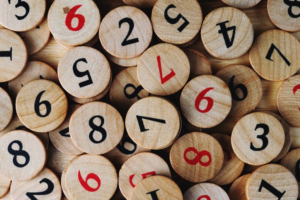 This image features a scattered collection of round wooden tokens, each printed with a single number. The discs vary slightly in tone, showing natural wood grain and texture. Numbers appear in both black and red ink, standing out clearly against the light brown surface. The tokens overlap and lie at different angles, creating a casual, tactile arrangement. Some numbers are upright while others are rotated, reinforcing the sense of randomness. The background appears to be a wooden tabletop, adding warmth and a handcrafted feel. The image suggests hands-on learning, play-based education, or early numeracy activities. The natural materials and simple design evoke traditional learning tools rather than digital ones, emphasizing sensory engagement through touch, sight, and arrangement. The scene feels approachable, practical, and well-suited to teaching basic counting or number recognition.