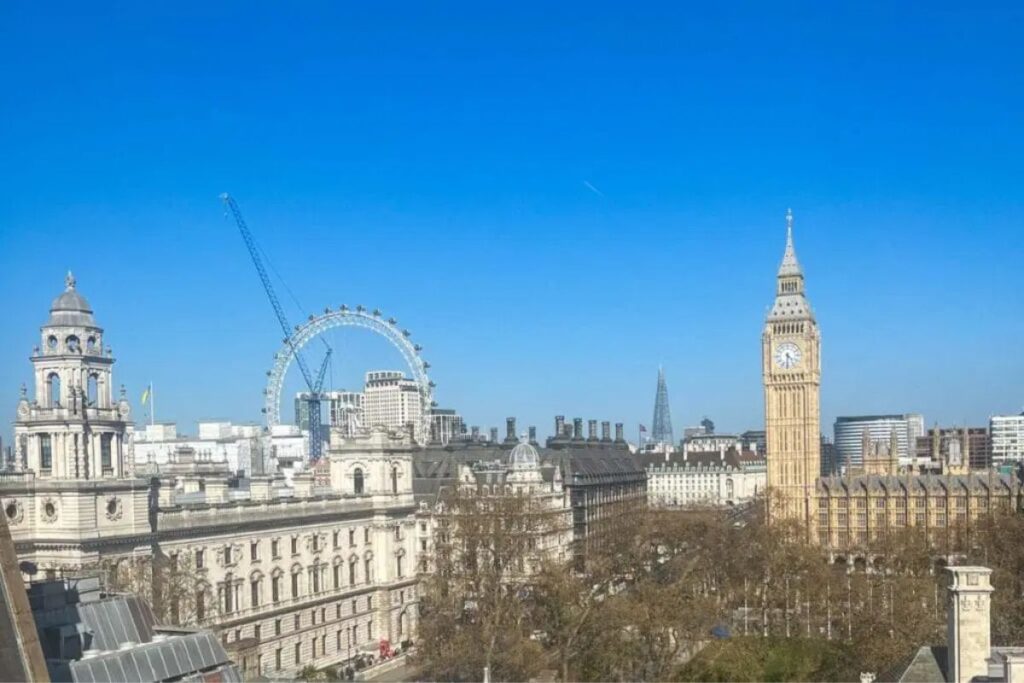 London Skyline This image presents a wide view of the London skyline under a vivid blue sky. Prominently featured on the right is the tall clock tower commonly known as Big Ben, rising above the surrounding buildings of the Palace of Westminster. To the left, the London Eye stands out with its large circular structure, partially framed by historic stone buildings and rooftops in the foreground. Bare trees fill the lower portion of the image, suggesting late winter or early spring. The mix of classical architecture and modern landmarks creates a layered cityscape, with rooftops, spires, and glass structures visible across the horizon. The clear weather and strong sunlight give the scene sharp detail and contrast, highlighting London’s blend of history, scale, and urban energy.