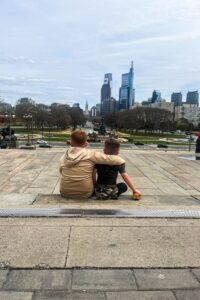 The image shows two children sitting side by side on wide stone steps, viewed from behind. One child has an arm wrapped around the other’s shoulders in a gentle, affectionate gesture. In front of them stretches a large urban park with pathways, trees, and a central monument. Beyond the park, a city skyline rises, featuring tall modern buildings and historic architecture in the distance. Cars move along the road below, and pedestrians are scattered throughout the scene. One child holds a small toy vehicle near their side, adding a quiet, playful detail. The image conveys a reflective pause during sightseeing, emphasizing companionship, scale, and the contrast between childhood closeness and the vast cityscape ahead.