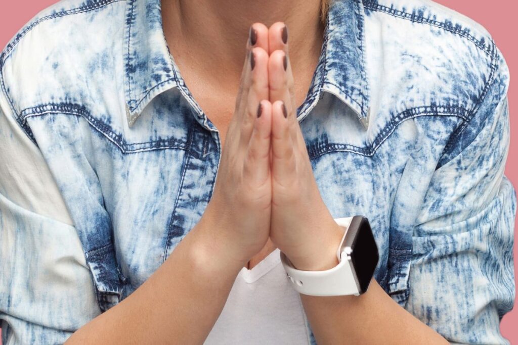 Say Please This image shows a person from the shoulders down with their hands pressed together in front of their chest, resembling a gesture of politeness, gratitude, or request. The individual wears a light denim shirt layered over a white top, and a smartwatch with a light-colored band is visible on one wrist. The background is a soft pink, providing a gentle contrast to the clothing. The posture and hand position suggest humility, respect, or asking politely. The absence of the person’s face keeps the focus on body language rather than identity, making the gesture universally relatable. The image visually communicates manners, empathy, and respectful interaction without the need for words.