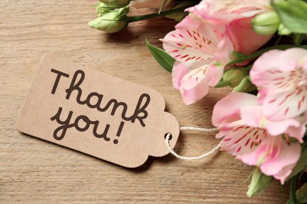 This image shows a close-up, warmly lit scene featuring a small wooden tag engraved with the words “Thank you” in a handwritten-style font. The tag is attached with a thin piece of twine and rests on a textured wooden tabletop, adding a natural, rustic feel. Beside the tag lies a delicate bouquet of soft pink flowers with pale green stems and buds, arranged loosely so that the petals gently frame the message. The flowers appear fresh and slightly open, suggesting care and thoughtfulness. The natural wood grain of both the tag and the surface beneath it creates a calm, earthy backdrop that enhances the sense of sincerity and appreciation. Overall, the image conveys gratitude, warmth, and kindness, making it well suited for themes related to saying thank you, expressing appreciation, or polite communication.