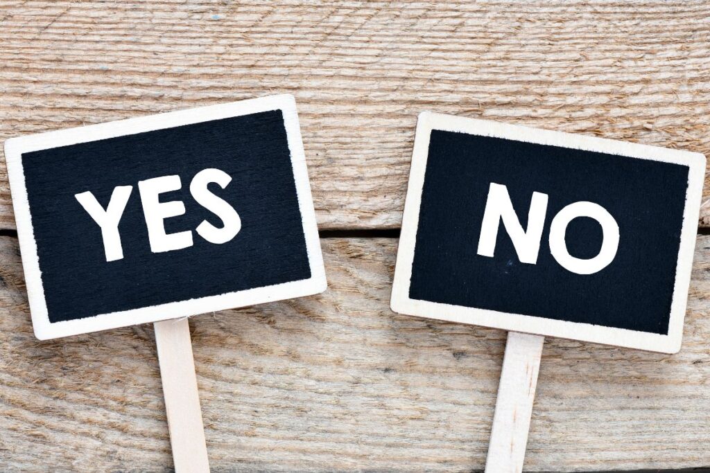 This image shows two small chalkboard-style signs placed on a wooden surface. One sign reads “YES” and the other reads “NO,” both written in bold white lettering. The signs are mounted on short wooden sticks and positioned slightly apart, emphasizing contrast and choice. The wood grain background adds a rustic texture, while the black-and-white signs create strong visual clarity. The composition highlights decision-making, boundaries, and contrasting responses. The image visually represents choice, consent, and the ability to decide between acceptance and refusal. It is often used in discussions about communication, assertiveness, and personal agency.