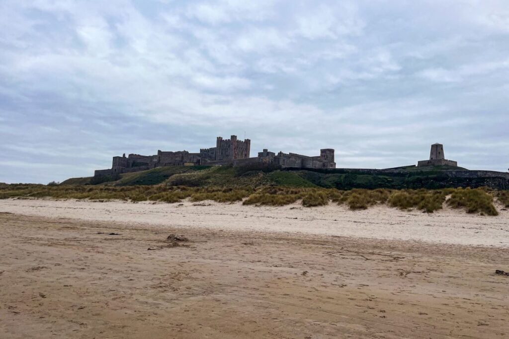 A wide sandy beach stretches across the foreground beneath an overcast sky, leading toward the historic stone walls of Bamburgh Castle in the distance. The large medieval fortress sits on a grassy ridge overlooking the coastline, with thick defensive walls and a central keep rising above the surrounding structures. The castle’s weathered stone blends into the muted tones of the cloudy sky, giving the scene a dramatic and slightly moody atmosphere. Low grassy dunes and patches of scrubby vegetation sit between the beach and the castle walls. To the right of the main castle complex stands a smaller stone tower structure on the hilltop. The beach itself appears quiet and mostly empty, with flat sand stretching toward the viewer. The overall image captures the imposing scale of Bamburgh Castle against the wide, open coastal landscape of Northumberland.