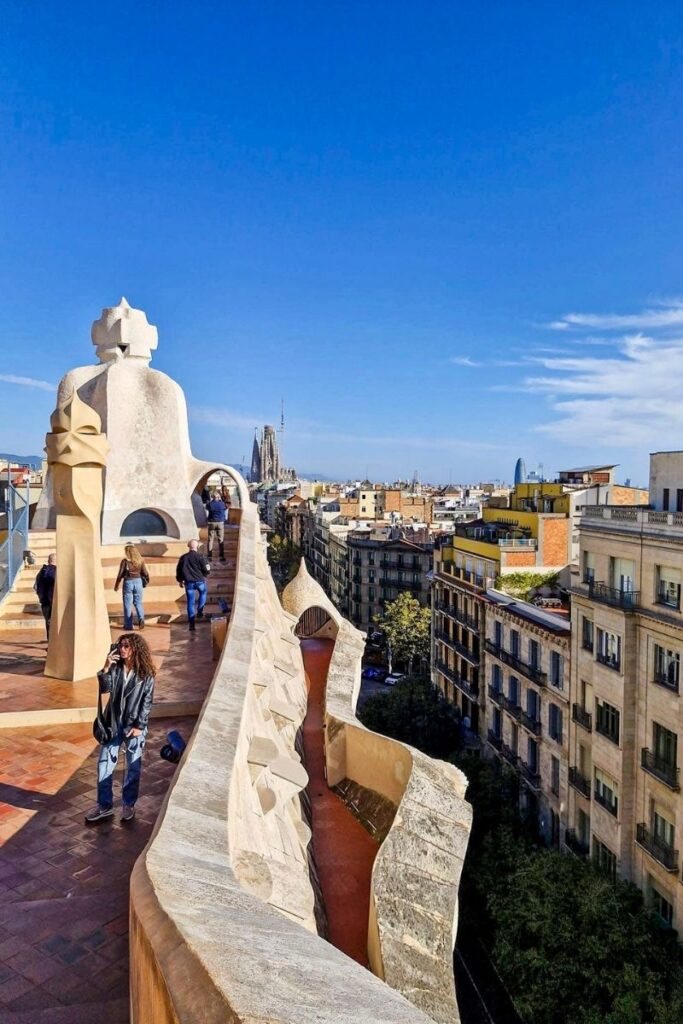 Barcelona The image shows a rooftop terrace in Barcelona with distinctive curved architectural features and pale stone structures typical of Antoni Gaudí’s design. The terrace has a warm terracotta-tiled floor and sculptural chimney-like forms with rounded edges and cut-out openings. Several people walk along the rooftop, pausing to take in the views. In the distance, the skyline stretches across the frame, with the spires of the Sagrada Família clearly visible rising above the surrounding buildings. To the right, rows of apartment buildings line a tree-filled street below. The sky is a vivid blue with only a few wispy clouds, and bright sunlight casts sharp shadows across the terrace. The image captures a blend of artistic architecture and sweeping city views from an elevated vantage point.