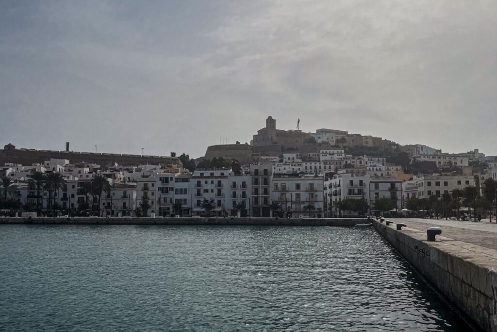Boat Trip Ibiza A waterfront view shows a coastal town built along the edge of a calm harbour. White buildings with flat roofs and balconies line the shoreline, reflecting traditional Mediterranean architecture. Higher up the hill behind the town sits an old stone fortress or historic structure overlooking the harbour. The water in the foreground ripples gently and reflects sunlight from the bright sky above. A long stone pier extends out along the right side of the image. Palm trees and waterfront walkways are visible among the buildings. The sky appears hazy with the sun shining through thin clouds, creating a soft glow over the town. The scene presents a peaceful seaside setting with historic architecture rising above the harbour.
