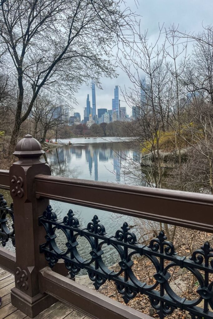 The view looks across a calm lake in Central Park toward the skyline of Manhattan in the distance. In the foreground, an ornate dark metal railing and wooden bridge frame the scene, suggesting the photo was taken from one of the park’s historic footbridges. Bare tree branches and early spring foliage surround the lake, creating a natural frame around the water. The surface of the lake is still enough to reflect the tall buildings of the city skyline beyond the park. Several modern skyscrapers rise above the trees, partially softened by light haze. The park landscape includes rocky outcrops and natural shoreline along the water’s edge. The contrast between the quiet greenery of Central Park and the dense urban skyline of New York City highlights how the park serves as a peaceful retreat within the busy city.