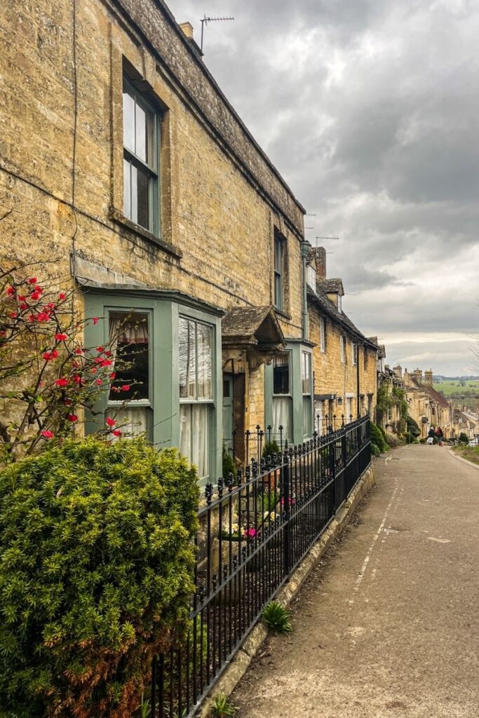 Cotswold Village Street A quiet street lined with traditional stone houses stretches downhill through a historic village. The buildings are constructed from warm yellow stone and feature tall windows, small doorways, and pitched roofs. Black wrought-iron railings run along the sidewalk in front of the houses. Small plants and shrubs grow in front gardens, including a bush with bright red flowers. The road gently slopes downward toward open countryside visible in the distance. Overhead, thick grey clouds cover the sky, creating a moody atmosphere. A few people walk further down the street near the bend in the road. The scene conveys the charm of an old English village, with preserved stone architecture and peaceful residential surroundings.