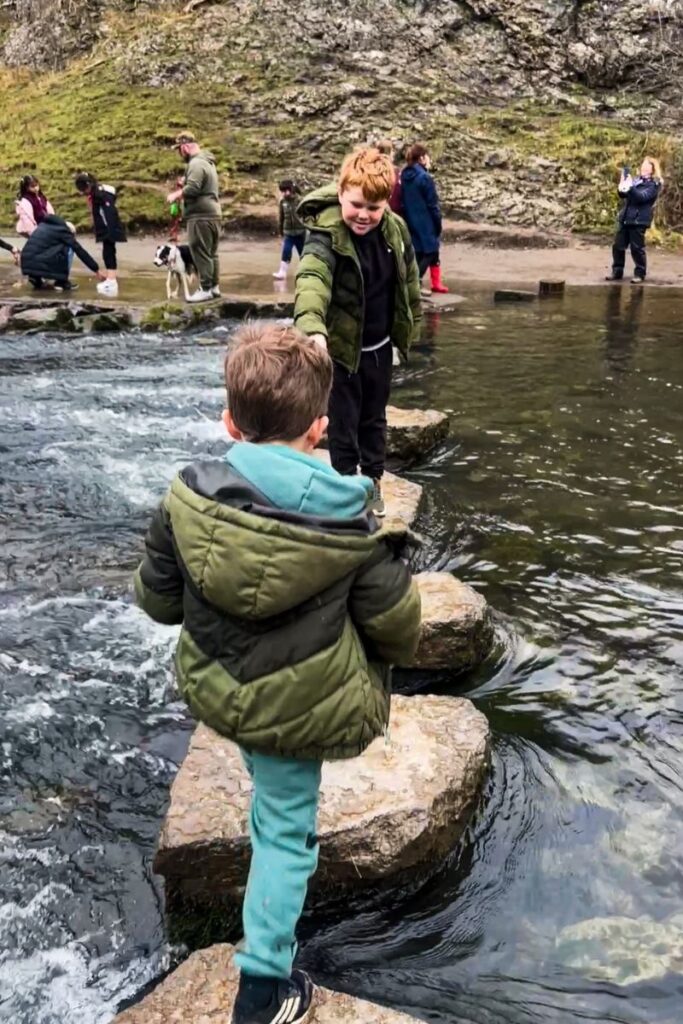 Dovedale Stepping Stones Two children are carefully crossing a shallow stream using large stepping stones placed across the water. The older child stands ahead, reaching back with one hand to help the younger child step onto the next stone. Both are wearing warm jackets, suggesting cool weather. The stream flows gently around the stones, creating small ripples and splashes. On the opposite bank, several adults and children stand watching or taking photos. A dog on a leash stands beside one of the adults. Behind them, rocky slopes and patches of grass form a natural landscape. The scene captures a moment of teamwork and outdoor adventure as the children balance across the stones while exploring the natural surroundings.
