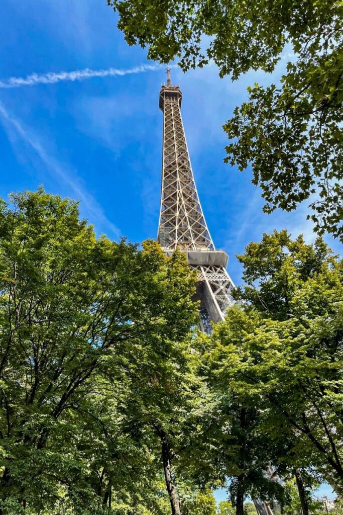 The Eiffel Tower rises dramatically into a bright blue sky, viewed from below and framed by green tree branches on either side. The famous iron lattice structure narrows as it rises upward, emphasizing its height and intricate metal framework. Sunlight illuminates the tower, highlighting the detailed cross-bracing of the steel beams. A thin white contrail stretches diagonally across the sky behind the tower, adding a subtle line through the background. The tops of leafy trees fill the lower portion of the image, partially surrounding the base of the structure and creating a natural frame. The perspective from below makes the tower appear especially tall and impressive as it towers above the trees. The scene captures the iconic landmark in a bright daytime setting, with clear weather and vibrant greenery adding contrast to the tower’s iron structure.
