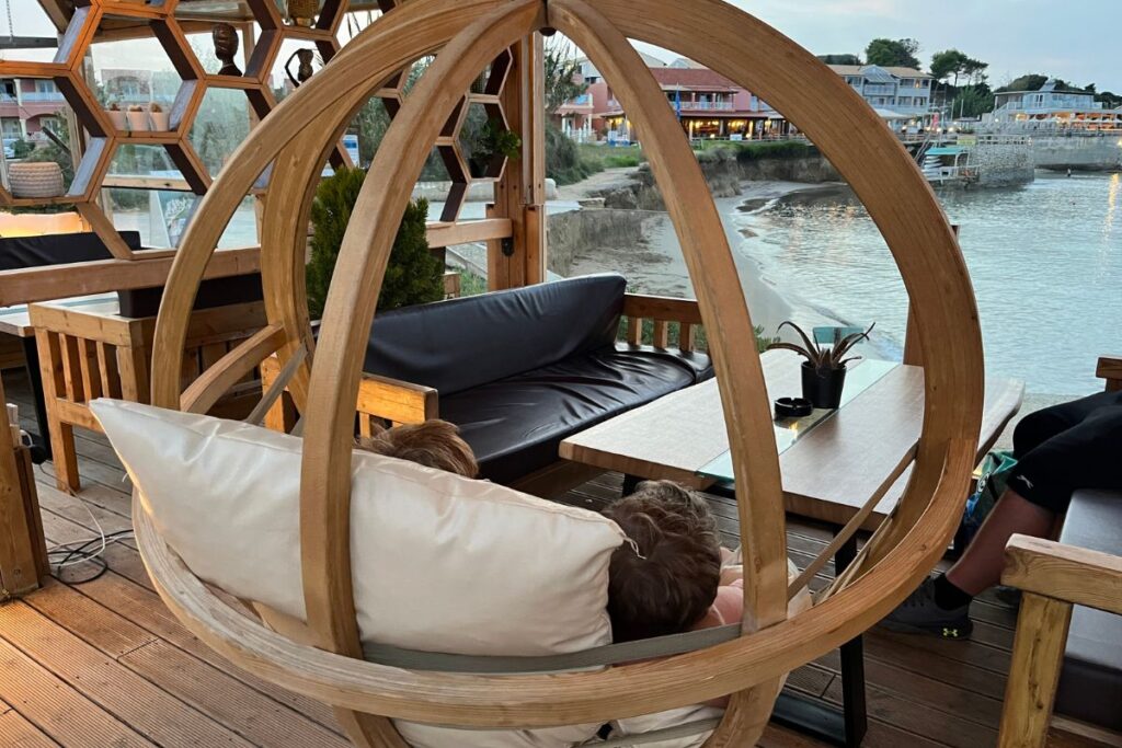 The image shows two children resting together inside a large, circular wooden hanging chair suspended from the ceiling of a covered outdoor seating area. The chair is shaped like an open ring and filled with light-colored cushions, creating a cozy, nest-like space. The children lie back comfortably, facing away from the camera, appearing relaxed and calm. In front of them is a low wooden table and a dark cushioned bench, suggesting a café or lounge setting. Beyond the seating area, the view opens onto a quiet shoreline with water gently meeting a sandy edge. Buildings with balconies and warm lights line the opposite side of the water, indicating a coastal town. The overall atmosphere feels peaceful and intimate, blending comfort with scenic seaside surroundings.
