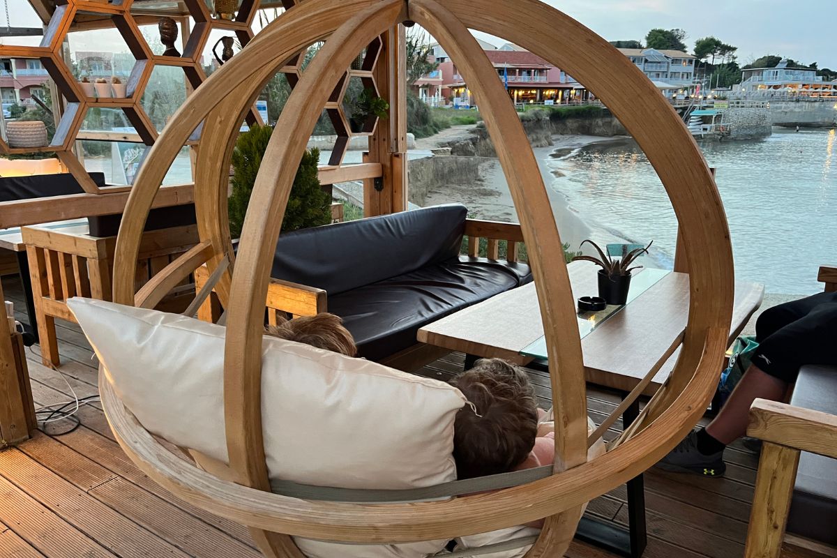 The image shows two children resting together inside a large, circular wooden hanging chair suspended from the ceiling of a covered outdoor seating area. The chair is shaped like an open ring and filled with light-colored cushions, creating a cozy, nest-like space. The children lie back comfortably, facing away from the camera, appearing relaxed and calm. In front of them is a low wooden table and a dark cushioned bench, suggesting a café or lounge setting. Beyond the seating area, the view opens onto a quiet shoreline with water gently meeting a sandy edge. Buildings with balconies and warm lights line the opposite side of the water, indicating a coastal town. The overall atmosphere feels peaceful and intimate, blending comfort with scenic seaside surroundings.