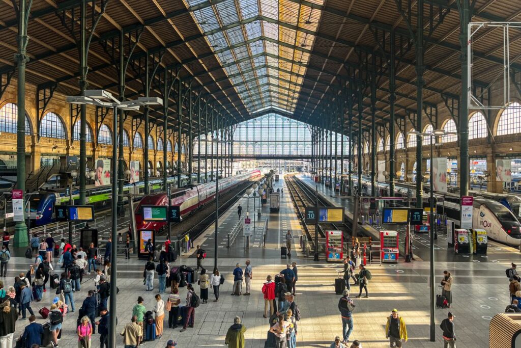 Gare Du Nord View The interior of a grand railway station stretches out beneath a high arched glass and steel roof. Multiple train platforms run parallel across the station floor, each marked with numbered signs. Several trains sit alongside the platforms, including sleek high-speed trains and regional trains in different colours. Passengers walk through the station carrying luggage, standing in small groups while waiting for departures. Sunlight filters through the glass roof, casting bright patches of light across the polished floor. Tall iron columns support the structure, giving the station a historic industrial style. Information boards, ticket machines, and platform signs are scattered throughout the space. The scene conveys the busy atmosphere of a major transport hub connecting different destinations.