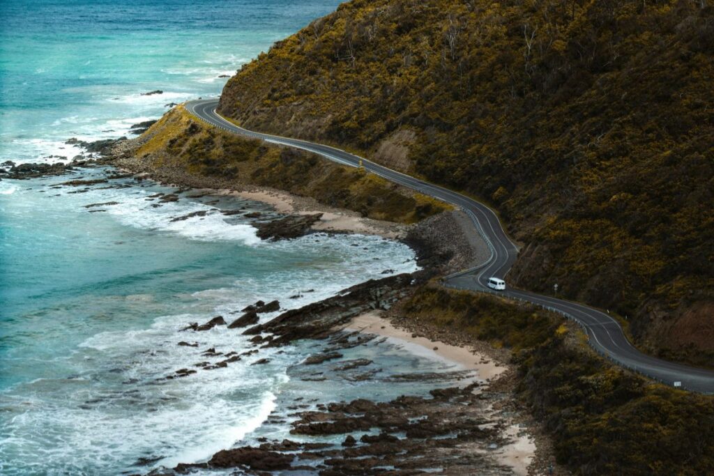 A winding coastal road curves dramatically along the rugged cliffs of the Great Ocean Road in Australia. The narrow asphalt road hugs the edge of the coastline as it snakes around a hillside covered in dry shrubs and vegetation. A small white van drives along one of the bends, giving a sense of scale to the landscape. To the left of the road, turquoise ocean waves roll toward the rocky shoreline, breaking into white foam as they hit the rocks and small sandy coves below. Dark rock formations and tide pools are scattered along the coast where the sea meets the land. The hillside rises steeply above the road, covered in dense brown and green foliage. The scene captures the dramatic beauty of one of Australia’s most famous scenic drives, where the road winds between steep cliffs and the vast ocean.