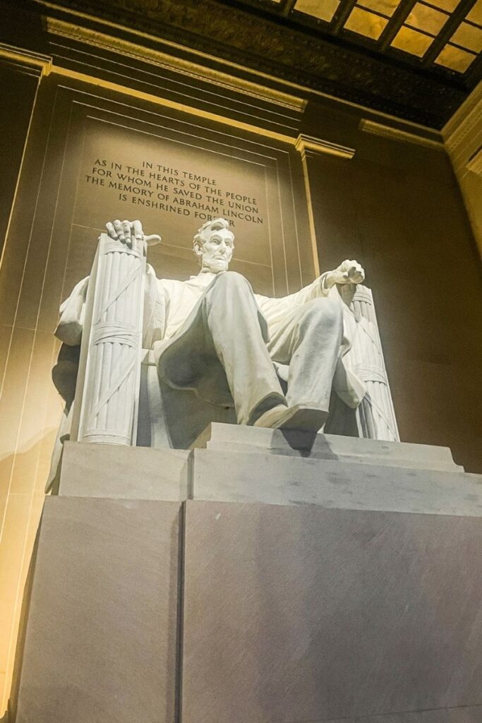 The massive white marble statue of Abraham Lincoln sits prominently inside the Lincoln Memorial in Washington, D.C. The seated figure of Lincoln rests in a large carved chair, with his hands placed firmly on the armrests. The statue is illuminated by warm interior lighting that highlights the detailed folds of the marble sculpture and Lincoln’s thoughtful expression. Behind the statue, a large stone wall bears an inscription that reads: “In this temple as in the hearts of the people for whom he saved the union the memory of Abraham Lincoln is enshrined forever.” Tall columns and a high ceiling frame the statue, emphasizing the scale and solemn atmosphere of the memorial. The perspective from below makes the statue appear even more imposing, reinforcing the sense of reverence associated with this historic monument.