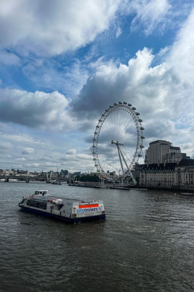 The River Thames flows through the foreground with a sightseeing boat traveling across the water. The boat is branded with “City Cruises” on the side and moves slowly along the river carrying passengers. In the background, the London Eye rises high above the riverbank, its large circular structure made of steel spokes and glass passenger capsules. The observation wheel stands beside several buildings along the South Bank of London. Above, large white and grey clouds fill the sky, creating a dramatic backdrop behind the London skyline. The river surface appears slightly rippled as the boat moves through the water. The scene captures a classic view of London, combining one of the city’s most recognizable landmarks with a tourist river cruise passing along the Thames.
