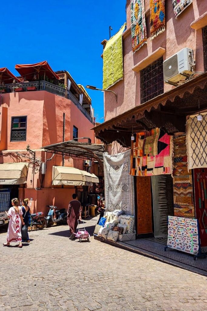 Marrakech This image captures a lively street in Marrakech lined with warm-toned buildings in shades of terracotta and peach. On the right side, a shop displays a variety of patterned rugs and textiles hanging vertically, featuring geometric designs in bright colours such as red, orange, cream, and black. Cushions are stacked at the entrance, adding texture and depth. A few people walk along the cobbled street, including one person pulling a small wheeled cart. On the left, a building with a shaded balcony and red umbrellas overlooks the street below. A scooter is parked near the centre, and shop awnings provide shade to nearby storefronts. The sky above is clear and intensely blue, enhancing the vibrant colours of the scene. The image conveys a busy yet relaxed market atmosphere rich in local character.