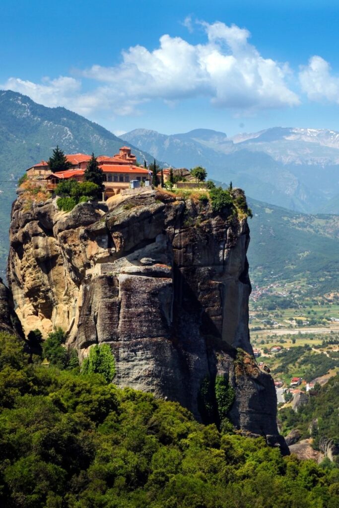 A dramatic view shows a historic monastery perched high on top of a towering rock pillar in Meteora, Greece. The monastery buildings have warm terracotta roof tiles and pale stone walls, contrasting with the dark grey vertical cliffs that rise sharply from the surrounding landscape. The rock formation appears almost column-like, with steep sides that drop down to dense green forest and shrubs at the base. A narrow stone staircase carved into the rock can be seen winding partway up the cliff toward the monastery. In the background, a wide valley stretches out with scattered houses, farmland, and winding roads, while layered mountain ranges fade into the distance. Above the scene, a bright blue sky with soft white clouds adds depth and scale. The image captures the extraordinary location of the Meteora monasteries, built atop natural rock formations overlooking the Thessalian plain.