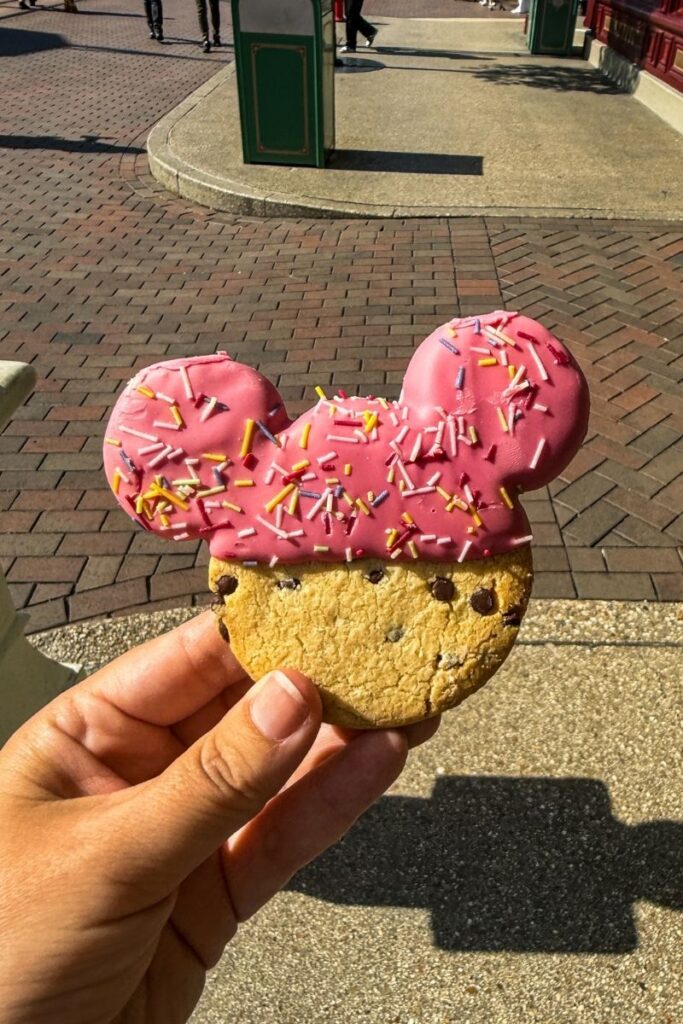 A hand holds a Mickey Mouse–shaped cookie in the foreground of the image. The cookie is designed with the recognizable silhouette of Mickey’s head and ears. The top portion is coated in bright pink icing and decorated with colorful sprinkles in shades of yellow, white, purple, and red. The lower portion of the cookie appears golden brown and slightly textured, with visible chocolate chips baked into the dough. The hand holding the cookie is positioned over a brick-paved walkway in an outdoor area that appears to be part of a theme park. In the background, green trash bins, shopfronts, and a few people walking around can be seen in soft focus. Bright sunlight casts strong shadows on the ground, emphasizing the cheerful and playful atmosphere associated with theme park snacks.