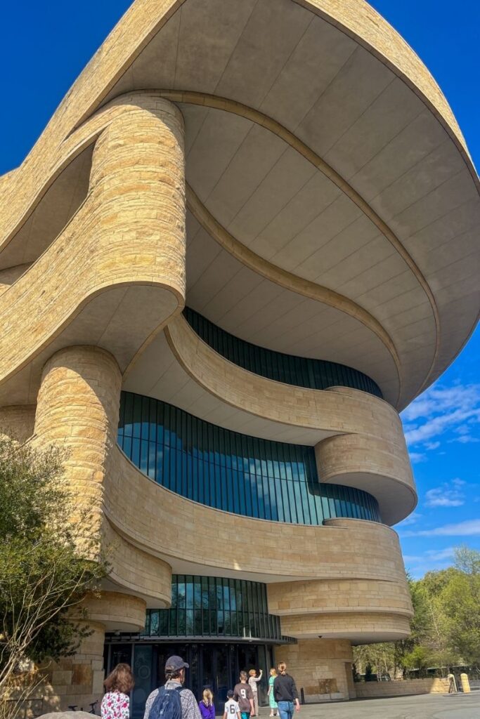 National Museum of the American Indian A large, modern building with sweeping curved architecture dominates the image. The structure is made of pale stone with layered, rounded balconies and cylindrical columns that support the upper levels. Large vertical windows with dark glass panels wrap around the middle floors, reflecting the bright blue sky above. The building’s flowing curves create an organic, sculptural appearance. At ground level, a small group of visitors, including adults and children, walk toward the entrance doors, providing a sense of scale compared to the massive structure. Trees and greenery frame the sides of the scene, and the bright sunlight casts soft shadows across the stone facade. The building appears to be a museum or cultural centre, designed with modern architectural style and sweeping lines.