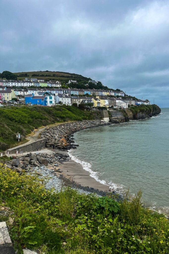 A picturesque seaside village stretches along the top of a rocky coastline overlooking the ocean. Rows of small houses painted in pastel colours—blue, yellow, pink, and white—line the cliff edge, creating a vibrant strip of colour against the grey sky. Below the houses, steep rocky slopes lead down to a narrow beach and a small curved bay where waves gently roll onto the shore. A stone path and coastal walkway wind along the cliffside. The sea appears calm with soft ripples reflecting the cloudy sky. Low green vegetation grows along the cliff edge in the foreground. The overall scene shows a charming coastal settlement perched above the water, combining dramatic natural scenery with colourful architecture.