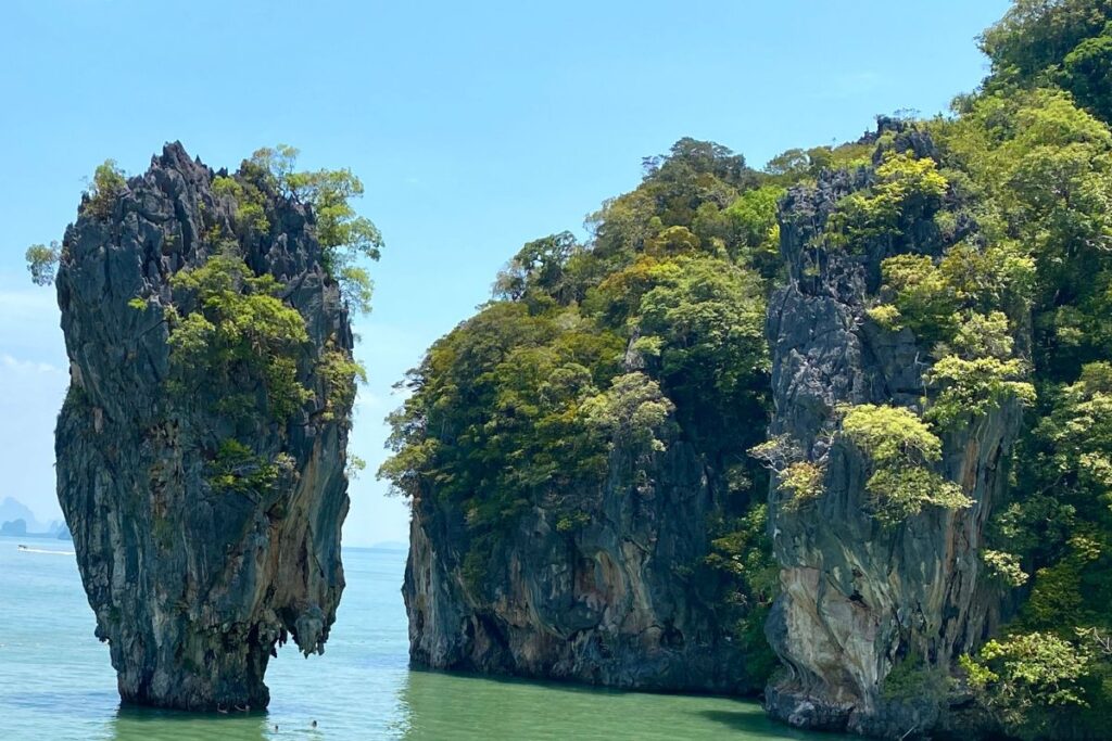 The image shows two towering limestone karst rock formations rising dramatically from calm green-blue water in Phang Nga Bay, Thailand. The tall rock pillar on the left stands almost vertically, with steep, jagged sides and small patches of vegetation clinging to the top and crevices in the rock. Beside it, a larger limestone cliff covered in dense green foliage slopes down toward the sea. The cliffs are textured with layers of grey and brown stone shaped by years of erosion. The water below appears shallow and tranquil, reflecting soft green tones from the surrounding landscape. In the distance, faint silhouettes of other islands can be seen across the horizon beneath a clear blue sky. The scene highlights the dramatic natural landscape of Phang Nga Bay, known for its striking limestone formations rising from tropical waters.