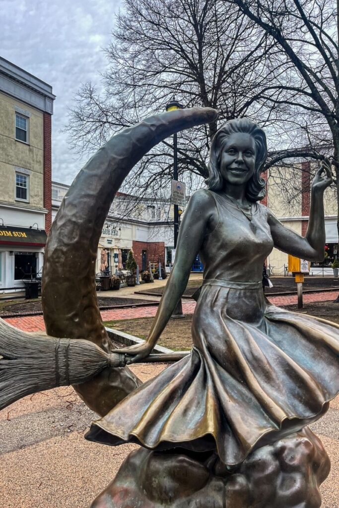 A bronze statue of a smiling woman dressed as a witch sits perched on a crescent moon while riding a broomstick. The sculpture depicts the witch wearing a flowing dress that flares outward as if caught in motion. One hand holds the broomstick while the other is raised in a playful gesture. The crescent moon curves behind her, forming part of the whimsical design of the statue. The bronze surface has a slightly weathered patina, reflecting years of outdoor display. In the background, a small town square with brick walkways, storefronts, and restaurants can be seen, along with leafless trees suggesting a cooler season. Signs for nearby shops are visible, placing the statue in a lively public area. The sculpture is a popular photo spot in Salem, Massachusetts, a town well known for its historical connection to witch trials.