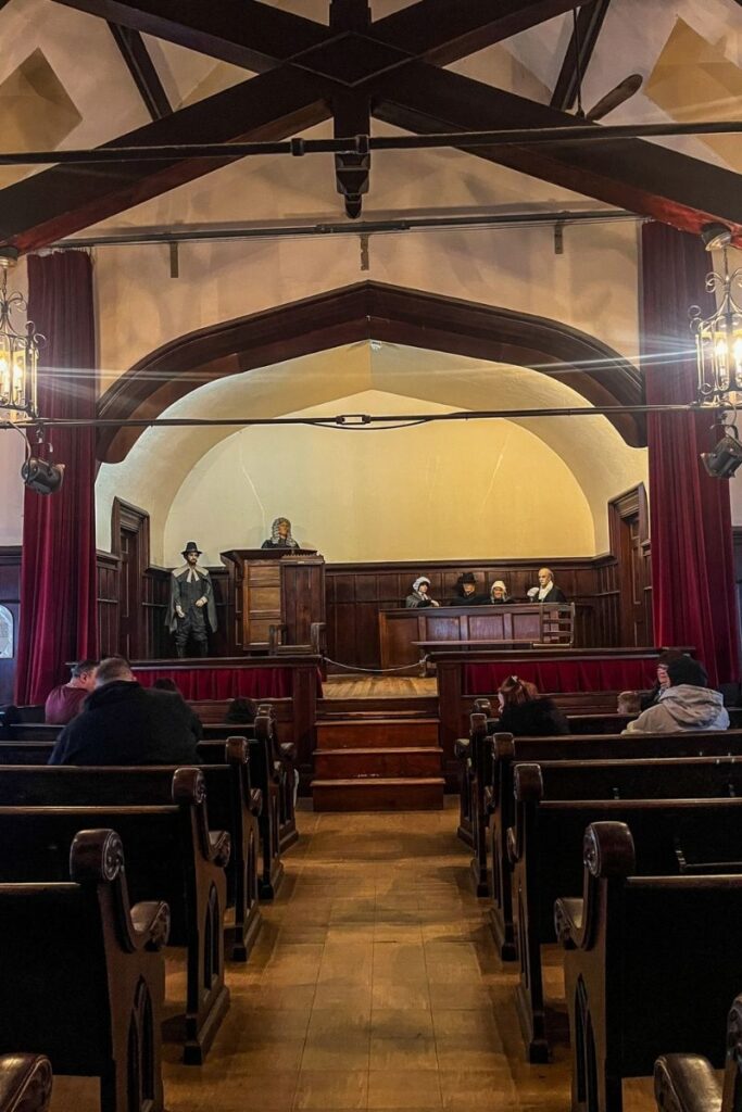 Salem Witch Performance The image shows the interior of an old courtroom designed with dark wooden paneling and traditional architecture. Rows of wooden benches fill the foreground where visitors sit facing the front of the room. At the far end, a raised wooden platform holds a judge’s bench and witness stand. Several life-sized mannequins dressed in historical clothing sit behind the bench, representing judges and court officials from earlier centuries. One figure stands near the witness stand wearing period attire including a hat and cloak. The ceiling features exposed wooden beams and two hanging lantern-style chandeliers that illuminate the room with warm light. Red curtains frame the stage-like courtroom area. The space resembles a preserved historical exhibit demonstrating what court proceedings may have looked like in the past.
