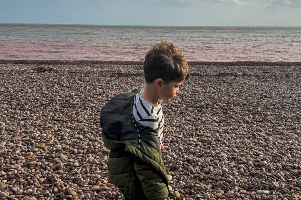 A young child stands near the edge of a wide pebble beach looking toward the sea. The child wears a green jacket, dark trousers, and trainers, with short hair slightly tousled by the breeze. Smooth round stones cover the entire beach, forming a textured foreground that stretches toward the water. The sea lies calm in the distance under a sky filled with layered clouds. Soft sunlight breaks through the clouds, illuminating the beach and creating a gentle glow on the water’s surface. The child stands slightly sideways, appearing thoughtful while looking toward the horizon. The scene feels quiet and reflective, capturing a peaceful moment beside the ocean on a cloudy day.