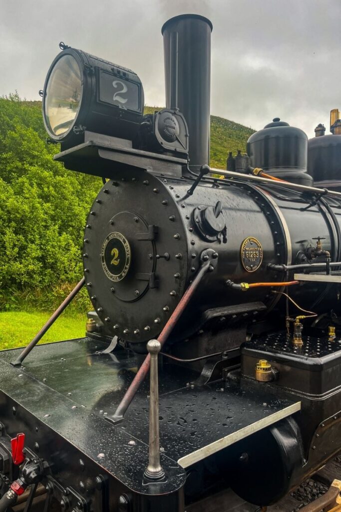 Steam Train in Wales A close-up view shows the front section of a historic black steam locomotive standing on railway tracks. The large cylindrical boiler dominates the image, with rivets and metal fittings clearly visible along its surface. At the front sits a round smokebox door bearing a circular metal plate marked with the number “2.” Above it, a large lantern-style headlamp with the number “2” displayed in white is mounted on a metal bracket. A tall black smokestack rises vertically from the top of the engine. Water droplets cover the metal surfaces, indicating recent rain. Behind the locomotive, green shrubs and grassy hills stretch into the distance under a cloudy sky. The scene captures the powerful, industrial design of early railway technology and the preserved condition of the historic engine.