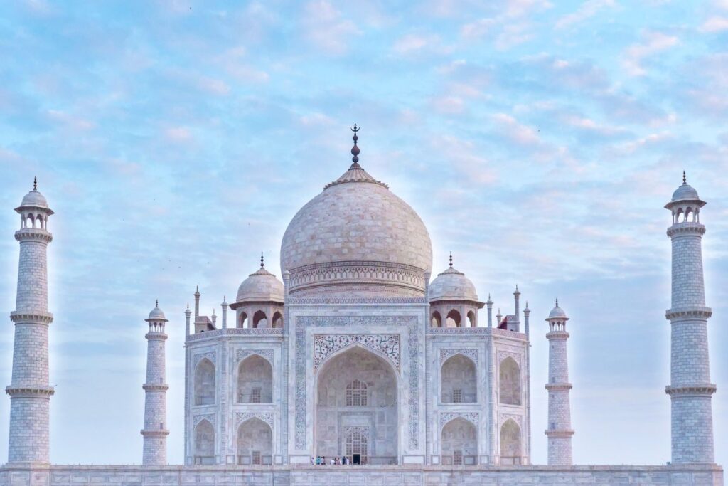 The Taj Mahal stands centered in the image, its large white marble dome rising above the ornate façade of the historic mausoleum in Agra, India. Four tall minarets stand symmetrically at the corners of the structure, framing the main building. The detailed marble surface is decorated with intricate carvings, arched doorways, and delicate patterns that reflect the architectural beauty of Mughal design. The central dome is topped with a decorative finial, while smaller domes surround it along the roofline. A soft pastel sky filled with thin clouds creates a gentle backdrop behind the monument. Small groups of visitors can be seen standing near the base of the structure, giving a sense of scale to the immense building. The image captures the balanced symmetry and elegance of one of the world’s most famous architectural landmarks.