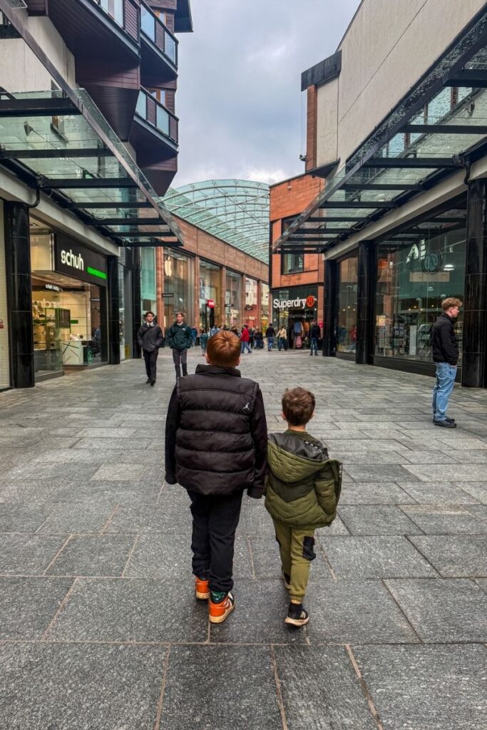 Walking City Tour Two children are walking away from the camera through a wide outdoor shopping area lined with modern storefronts. The older child, wearing a black padded vest, dark trousers, and orange trainers, walks beside a younger child dressed in a green jacket and matching trousers. They appear to be holding hands as they walk along a grey stone-paved pedestrian walkway. Glass canopies extend from the buildings on both sides, creating a sheltered corridor. Several shop signs are visible, including Schuh and Superdry. Other shoppers walk casually in the background, some entering and leaving stores. The buildings combine glass, brick, and metal architecture, and a curved glass roof structure spans the centre of the walkway further ahead. The sky above is overcast with dark grey clouds, suggesting cool or rainy weather.